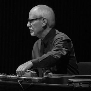 John Bischoff is seated behind various instruments and electronics. He looks out of frame to the left in profile.