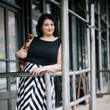 Wendy Richman holds her viola in her right arm as she leans against a wooden railing on a porch. She wears a black and white dress and smiles at the camera. 