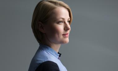 Portrait of a white woman with chin-length blonde hair, wearing a high necked light blue shirt. She is lightly smiling, nearly in side profile, with her face turned slightly  towards the camera.