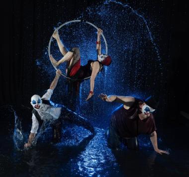 3 performer clowns, one on a cirque hoop, 2 reaching to the ground on blue lit water