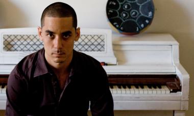 A portrait of a tan man with buzzed short hair wearing a dark maroon button up shirt is sitting at a white piano. He is in shadow, and stares at the camera solemnly but directly.