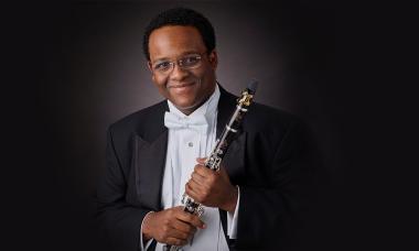 A Black man wearing a tuxedo with a white bow tie smiles at the camera. He is holding a clarinet.
