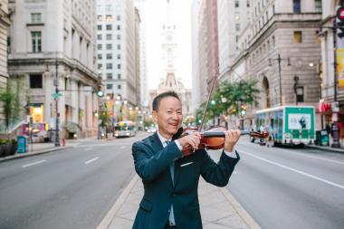 Soloist David Kim, holding a violin, standing on a meridian in the street, surrounded on both sides by buildings