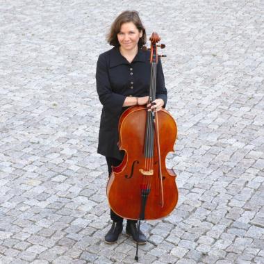 Natasha Jaffe standing and holding her cello on a grey cobblestone ground.