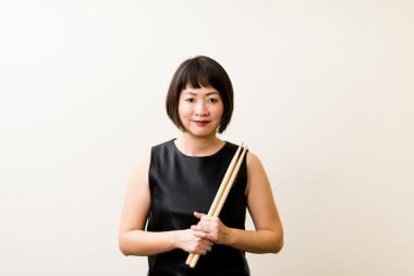Percussionist and composer Haruka Fujii stands in front of a white backdrop holding a pair of drumsticks.