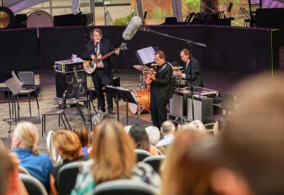 Members of the Brubeck Brothers Quartet performing bass, guitar, and drums in from of audience.