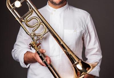 A bald man with light tan skin and a dark brown shadow of a beard, wearing glasses, a white button up shirt, and light jeans stares at the camera with a neutral expression. He holds a trombone with his right hand, his left in his pocket. 