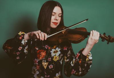 A white brunette feminine person with a blunt bob cut, red lipstick, wearing a floral long sleeve shirt, plays the violin.