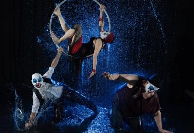 3 performer clowns, one on a cirque hoop, 2 reaching to the ground on blue lit water