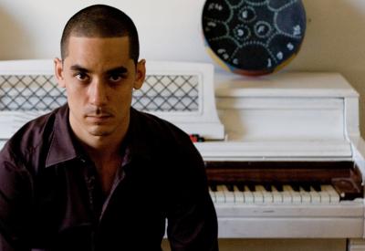 A portrait of a tan man with buzzed short hair wearing a dark maroon button up shirt is sitting at a white piano. He is in shadow, and stares at the camera solemnly but directly.