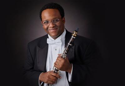A Black man wearing a tuxedo with a white bow tie smiles at the camera. He is holding a clarinet.