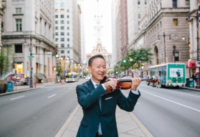 Soloist David Kim, holding a violin, standing on a meridian in the street, surrounded on both sides by buildings