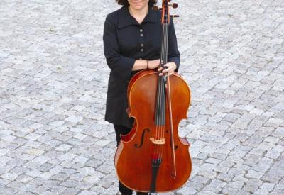 Natasha Jaffe standing and holding her cello on a grey cobblestone ground.