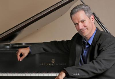 Pianist Robert Thies sits in front of a Steinway grand piano. The lid is open and the keys are uncovered. Thies sits with his back to it, his arm resting on the lifted fallboard.