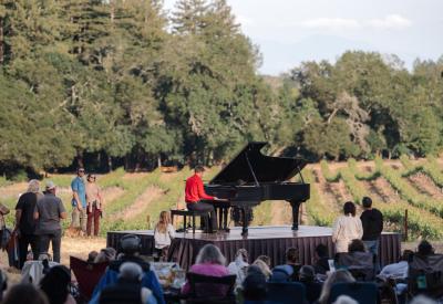 Hunter Noack plays piano in a field with audience members standing and seated in camping chairs while the sun sets