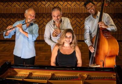 The Heloísa Fernandes Quartet stands smiling in front of a piano, each holding their instruments 